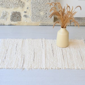 May include: A white woven rug with a fringed edge. The rug has a subtle striped pattern and is placed on a light gray floor. A vase with dried grass is in the background.