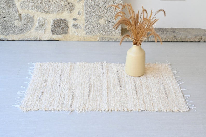 May include: A white woven rug with a fringed edge. The rug has a subtle striped pattern and is placed on a light gray floor. A vase with dried grass is in the background.