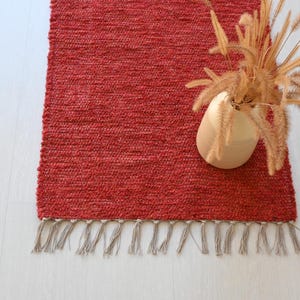 Peut inclure: Un tapis rectangulaire rouge tissé avec une bordure à franges. Un vase beige avec de l'herbe ornementale séchée est posé sur le tapis. Le tapis est sur un sol en bois clair.