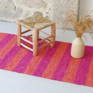 May include: A pink and orange striped rug with a woven texture. A small wooden stool with a woven seat sits on the rug. A vase with dried grass sits on the rug.