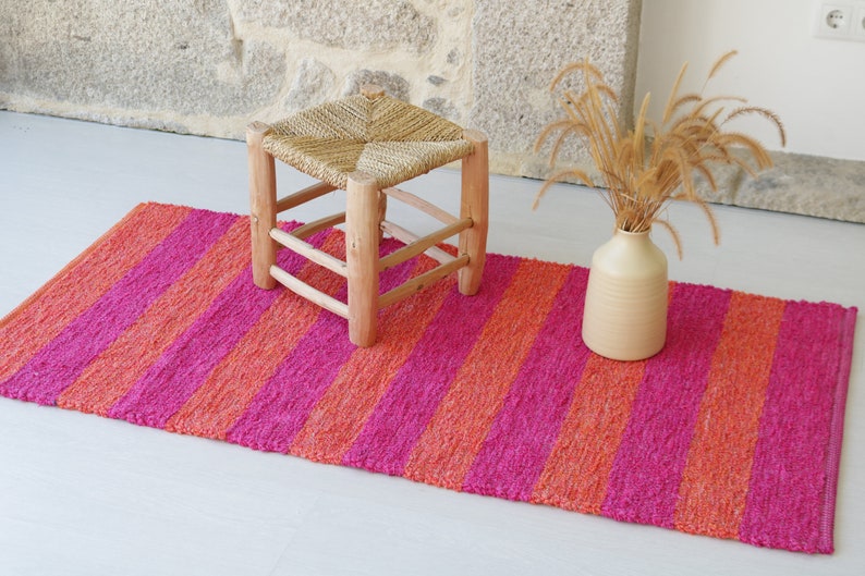 May include: A pink and orange striped rug with a woven texture. A small wooden stool with a woven seat sits on the rug. A vase with dried grass sits on the rug.