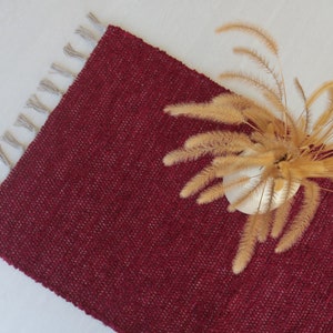 May include: A burgundy red woven rug with fringe on the edges. The rug is textured and has a subtle sheen. A dried flower arrangement is on top of the rug.