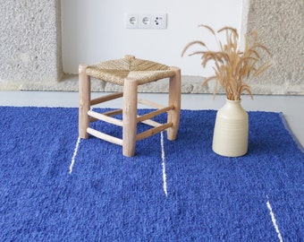 May include: A blue and white woven rug with a wooden stool and a vase of dried grass.