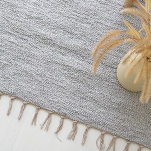May include: A light gray rug with a fringe border. The rug is made of a soft, textured material. A dried plant in a beige vase is placed on the rug.