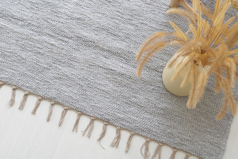 May include: A light gray rug with a fringe border. The rug is made of a soft, textured material. A dried plant in a beige vase is placed on the rug.