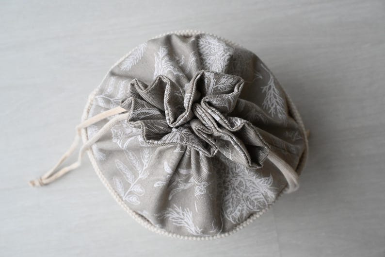 Pu&ograve; includere: Un sacchetto rotondo in tessuto grigio con chiusura a coulisse. Il sacchetto &egrave; decorato con un motivo botanico bianco e una parte superiore arricciata. Il cordino &egrave; di colore beige naturale.