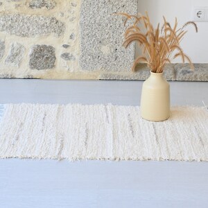 May include: A white woven rug with a fringed edge, placed on a light gray floor. A vase with dried grass is in the background.