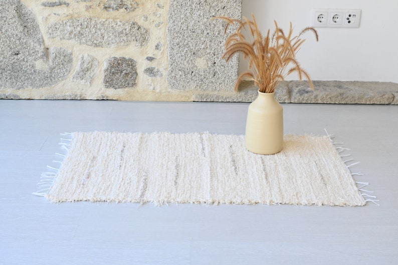 May include: A white woven rug with a fringed edge, placed on a light gray floor. A vase with dried grass is in the background.