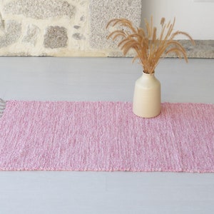 Puede incluir: Una alfombra tejida rosa con flecos en los extremos. La alfombra está sobre un suelo blanco y un jarrón con hierba seca está en el fondo.