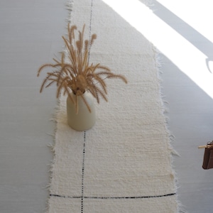 May include: A white fluffy rug with black lines forming a grid pattern. The rug has frayed edges and is placed on a light gray tiled floor. A vase with dried brown grass is centered on the rug.