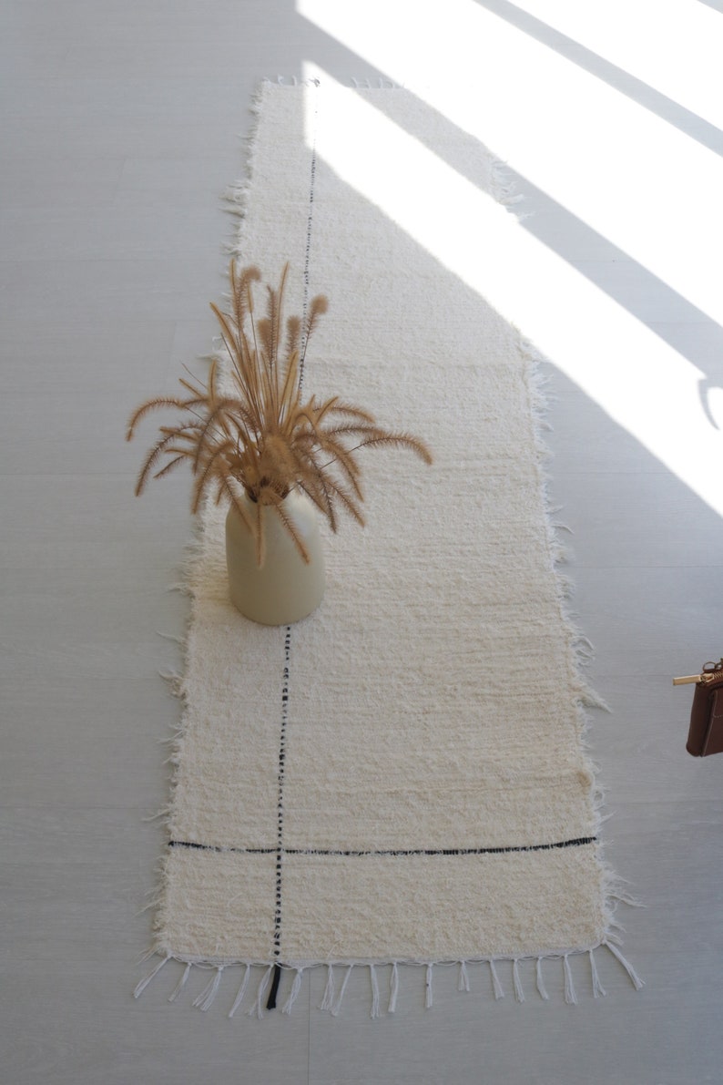 May include: A white fluffy rug with black lines forming a grid pattern. The rug has frayed edges and is placed on a light gray tiled floor. A vase with dried brown grass is centered on the rug.