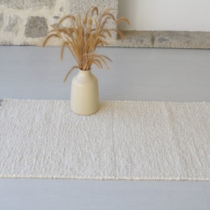 Puede incluir: Una alfombra blanca tejida con flecos en los bordes. La alfombra est&aacute; sobre un suelo gris claro y un jarr&oacute;n con hierba seca est&aacute; en el centro de la alfombra.