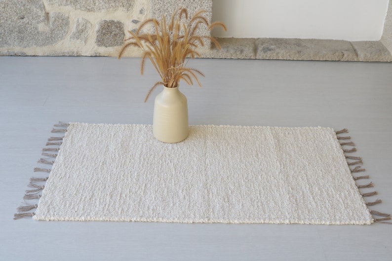 Puede incluir: Una alfombra blanca tejida con flecos en los bordes. La alfombra est&aacute; sobre un suelo gris claro y un jarr&oacute;n con hierba seca est&aacute; en el centro de la alfombra.