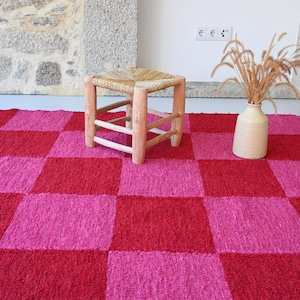 Pode incluir: Um tapete xadrez rosa e vermelho com textura tecida. Um banquinho de madeira com assento tecido está sobre o tapete. Um vaso com grama seca está sobre o tapete.