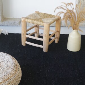 May include: A black woven rug with a woven stool and a vase with dried grass. The stool is made of wood and has a woven seat.