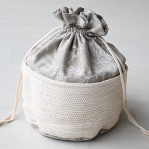 Pu&ograve; includere: Un cestino portaoggetti rotondo bicolore. L'esterno &egrave; in materiale tessuto bianco sporco, mentre l'interno presenta un tessuto grigio con un motivo floreale bianco. Una chiusura con coulisse assicura la parte superiore.