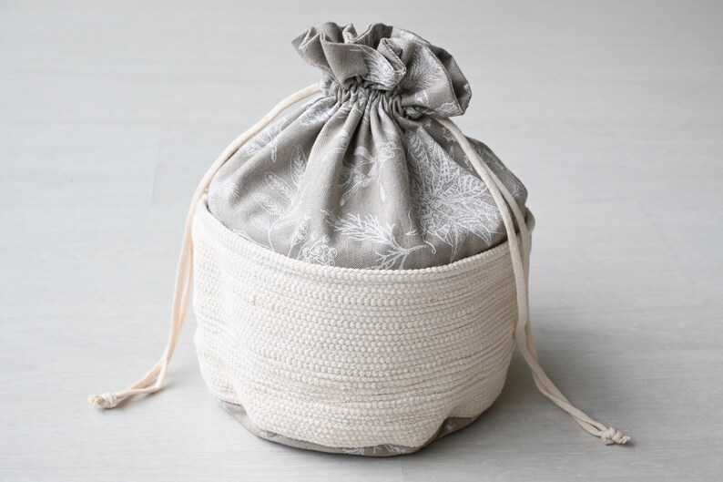 Pu&ograve; includere: Un cestino portaoggetti rotondo bicolore. L'esterno &egrave; in materiale tessuto bianco sporco, mentre l'interno presenta un tessuto grigio con un motivo floreale bianco. Una chiusura con coulisse assicura la parte superiore.