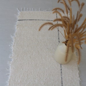 May include: A white, fluffy rug with a black line running across it. A vase with dried, tan grasses is on the rug.