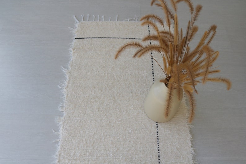 May include: A white, fluffy rug with a black line running across it. A vase with dried, tan grasses is on the rug.