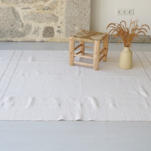 Puede incluir: Una alfombra blanca tejida con flecos en los bordes y dos sutiles rayas que recorren el centro. Un taburete de madera con un asiento tejido se encuentra sobre la alfombra, y un jarr&oacute;n con hierba seca se encuentra sobre la alfombra.