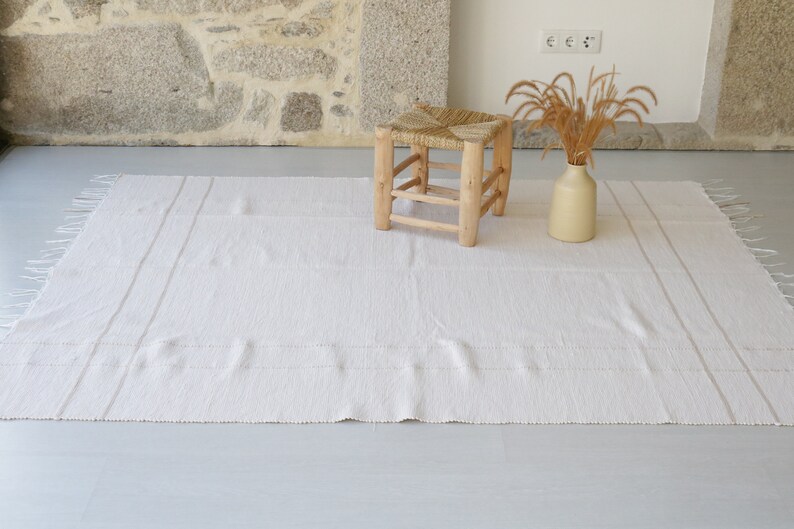 Puede incluir: Una alfombra blanca tejida con flecos en los bordes y dos sutiles rayas que recorren el centro. Un taburete de madera con un asiento tejido se encuentra sobre la alfombra, y un jarr&oacute;n con hierba seca se encuentra sobre la alfombra.