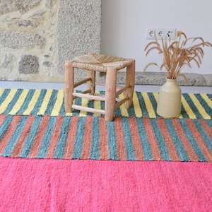 Peut inclure: Un tapis rectangulaire présentant des bandes horizontales de couleur, incluant une section rose vif unie, une section rayée avec des lignes alternées bleu sarcelle et terre cuite, et une section rayée avec des lignes jaune et bleu sarcelle.