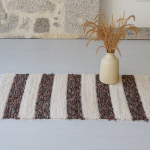 May include: A striped rug with cream and dark brown vertical bands. The rug has a fringed edge and is on a light grey surface. A beige vase with dried grass is behind the rug. The rug's dimensions are not visible.