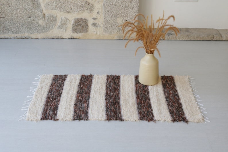 May include: A striped rug with cream and dark brown vertical bands. The rug has a fringed edge and is on a light grey surface. A beige vase with dried grass is behind the rug. The rug's dimensions are not visible.