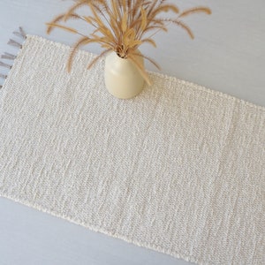 Puede incluir: Una alfombra tejida de color crema con flecos en todos los lados. La alfombra est&aacute; hecha de fibras naturales y tiene una textura sutil.