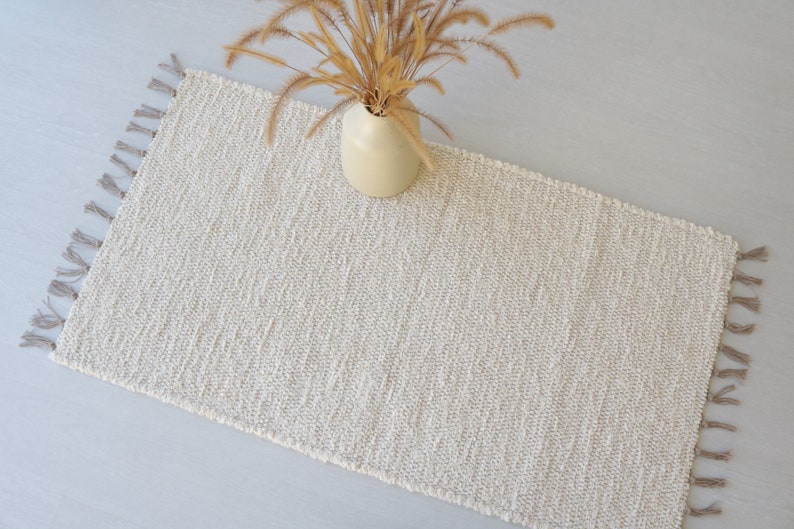 Puede incluir: Una alfombra tejida de color crema con flecos en todos los lados. La alfombra est&aacute; hecha de fibras naturales y tiene una textura sutil.