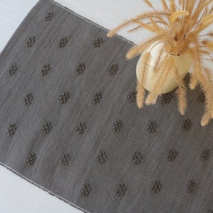 Può includere: Un tappeto tessuto grigio con un motivo ripetitivo di piccoli cerchi grigio scuro. Il tappeto è steso su una superficie bianca.