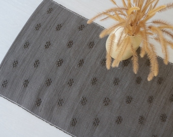 Puede incluir: Una alfombra tejida gris con un patrón repetitivo de pequeños círculos gris oscuro. La alfombra está tendida sobre una superficie blanca.