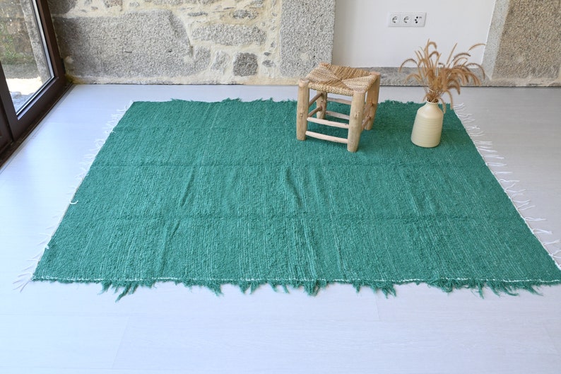 May include: A teal green woven rug with a fringed edge. A small wooden stool sits on the rug.