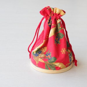 Pode incluir: Uma bolsa de tecido vermelha com cord&atilde;o de ajuste e um padr&atilde;o floral dourado e azul. A bolsa est&aacute; sobre uma base de madeira.