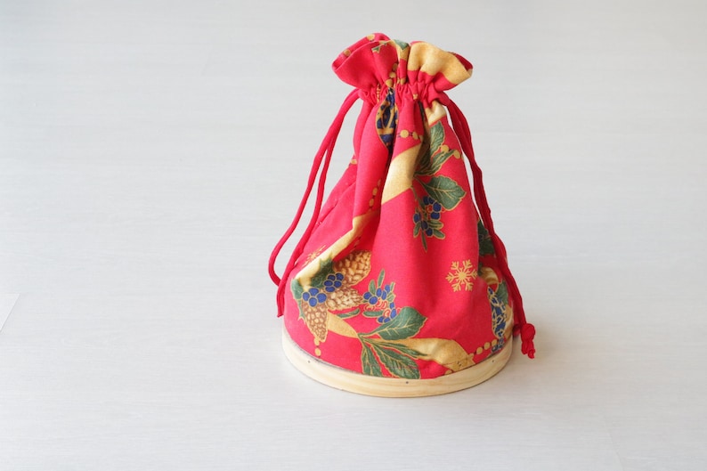 Pode incluir: Uma bolsa de tecido vermelha com cord&atilde;o de ajuste e um padr&atilde;o floral dourado e azul. A bolsa est&aacute; sobre uma base de madeira.