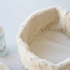 May include: A white, round, fabric-lined basket with a white background. A blue and white bottle of "Beb&eacute;s Mam&aacute;" baby lotion is visible in the foreground.