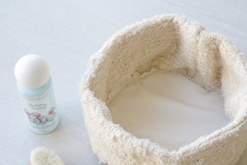 May include: A white, round, fabric-lined basket with a white background. A blue and white bottle of "Beb&eacute;s Mam&aacute;" baby lotion is visible in the foreground.