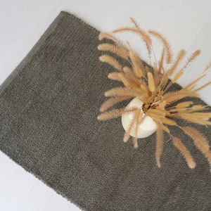 May include: A dark gray woven rug with a vase of dried pampas grass on top.