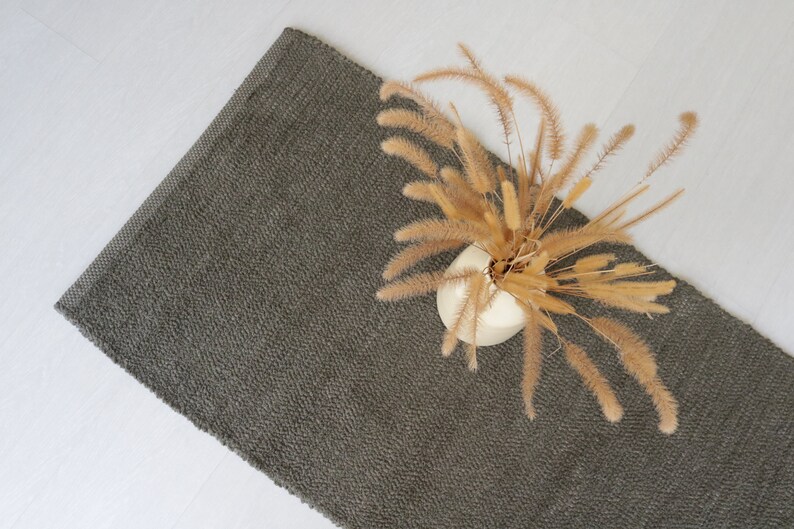 May include: A dark gray woven rug with a vase of dried pampas grass on top.