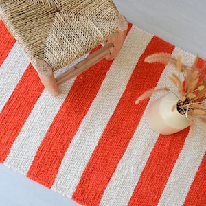 May include: A rectangular woven rug featuring alternating vertical stripes in bright orange and off-white. The rug has a textured, chunky knit appearance. A small wooden stool with a woven seat and a vase with dried grasses sit on the rug.
