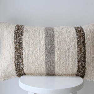 Pode incluir: Uma almofada branca com listras marrons e cinzas. A almofada é feita de um tecido macio e texturizado e tem uma borda ligeiramente desfiada.