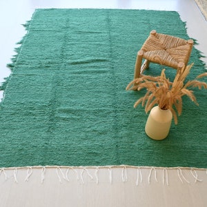 May include: A green woven rug with white fringe. The rug is on a light wood floor and has a small wooden stool and a vase of dried flowers on top of it.