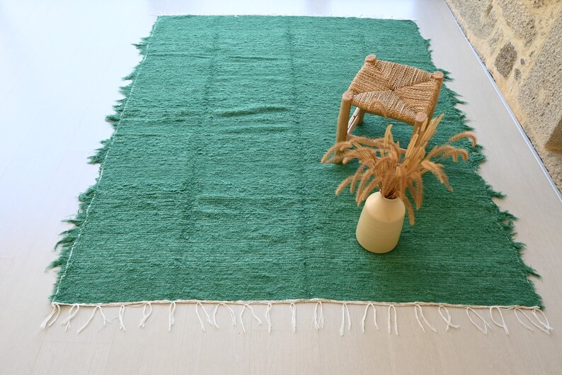 May include: A green woven rug with white fringe. The rug is on a light wood floor and has a small wooden stool and a vase of dried flowers on top of it.