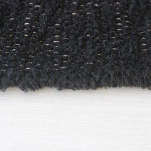 May include: Close-up of a black, textured fabric with a fringe edge. The fabric has a woven pattern with a subtle sheen.