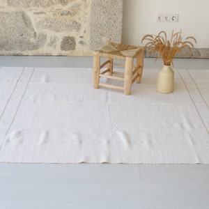 Puede incluir: Una alfombra blanca tejida con bordes con flecos y una textura sutil. Un taburete de madera con un asiento tejido se encuentra sobre la alfombra, y un jarr&oacute;n con hierbas secas est&aacute; en el suelo.