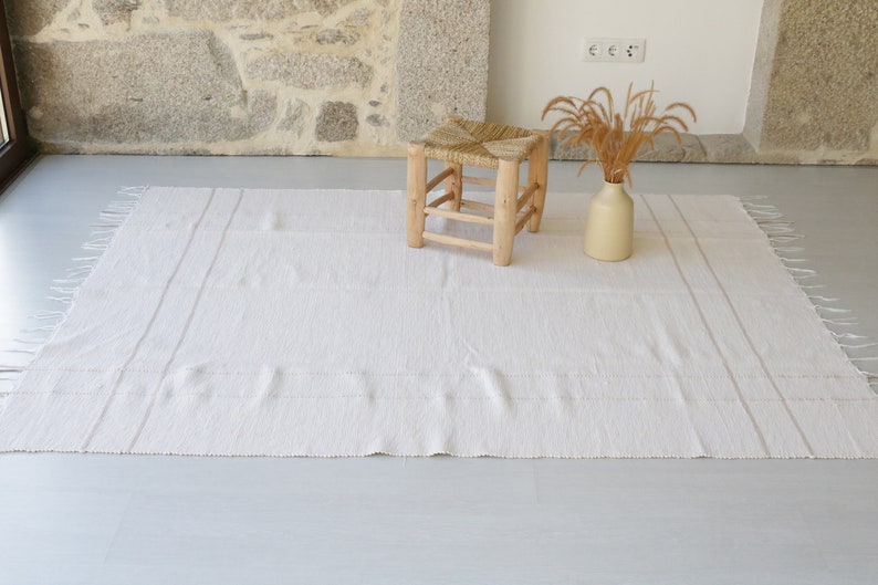 Puede incluir: Una alfombra blanca tejida con bordes con flecos y una textura sutil. Un taburete de madera con un asiento tejido se encuentra sobre la alfombra, y un jarr&oacute;n con hierbas secas est&aacute; en el suelo.