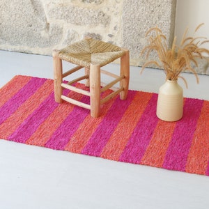 May include: A pink and orange striped rug with a wooden stool and a vase with dried grass.