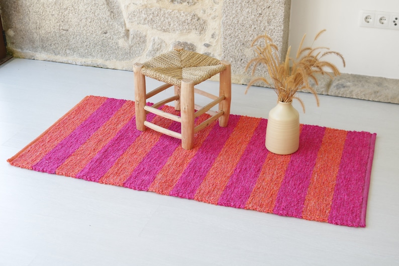 May include: A pink and orange striped rug with a wooden stool and a vase with dried grass.