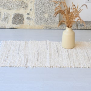 May include: A white woven rug with a fringed edge. The rug is laid out on a light gray floor with a vase of dried grasses in the background.