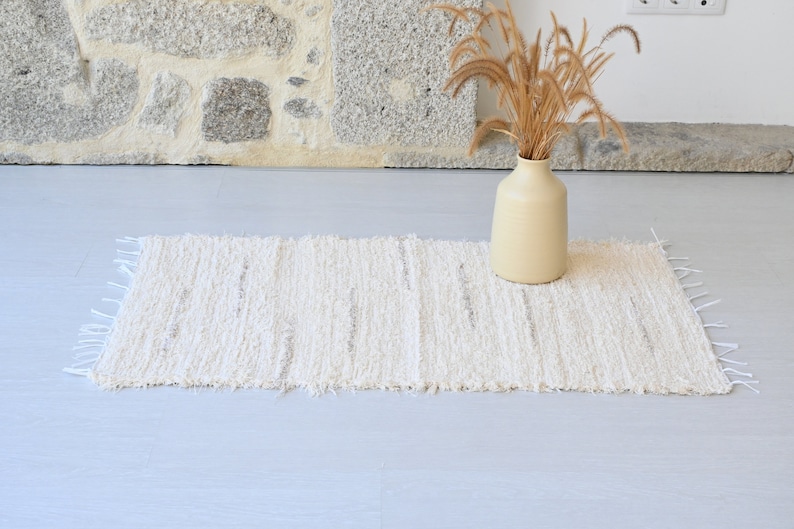May include: A white woven rug with a fringed edge. The rug is laid out on a light gray floor with a vase of dried grasses in the background.